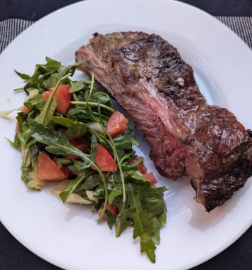 Bir parça pişmiş et, az pişmiş ve beyaz tabakta roka salatası. Koyu mavi arkaplan