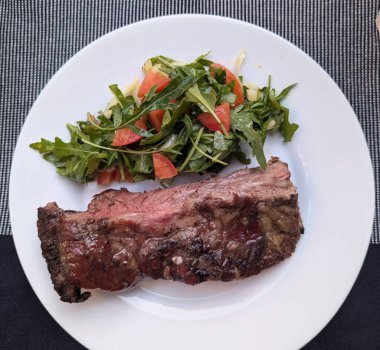 Bir parça pişmiş et, az pişmiş ve beyaz tabakta roka salatası.