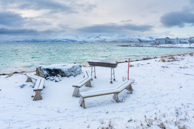 Karlı şömine ve kışın Kuzey Atlantik kıyısında ahşap park bankları. Hillesy ve Sommary adalarında arka planda ahşap yazlıklar. Troms, Kuzey Norveç 'te Tatil.