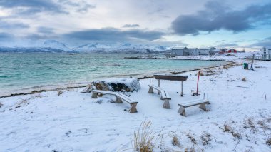 Karlı şömine ve kışın Kuzey Atlantik kıyısında ahşap park bankları. Hillesy ve Sommary adalarında arka planda ahşap yazlıklar. Troms, Kuzey Norveç 'te Tatil.