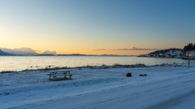 Karlı şömine ve kışın Kuzey Atlantik kıyısında ahşap park bankları. Hillesy ve Sommary adalarında arka planda ahşap yazlıklar. Troms, Kuzey Norveç 'te Tatil.