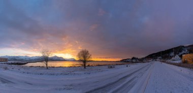 Norveç, Senja 'nın karlı dağları üzerinde parlak bir güneş ışığı parlıyor. Köprü ve Barbekü alanı, öndeki dinlenme alanı. Kırmızı bulutlar Skaland 'daki fiyordun suyuna yansıyor. Bergsfjorden 'da gün batımı Bergsoyan manzaralı