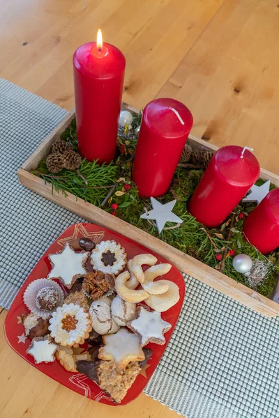 first advent candle burn on the advent decoration, placed by a boule filled with cookies. advent cookies on a red plate. modern advent wreath on the table
