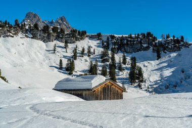 Ahşap dağ kulübesi, çatısı karlı, ağaçları ve taze karla kaplı dağları olan bir manzara. Karla kaplı çayırların ve ormanların üzerindeki dik uçurum kulesiyle dağlara çarpıyor.