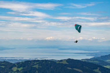 Mavi bulutlu gökyüzünde paraşüt broşürleri, yeşil çayır, köknar ağacı, mavi su ve dağ sırasıyla Constance Gölü üzerinde paraşüt manzarası. Bregenz Ormanı, Vorarlberg 'de yaz günü,