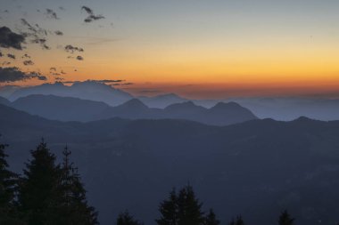 Bregenzerwald panoramasında dağ siluetleri, arka planda İsviçre dağları. Gün batımı kırmızı ve sisli hatları görünür kılar..