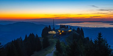 Akşam vakti Bezau Baumgarten teleferiğinin aydınlık dağ istasyonu, dağların üzerinde gün batımı, arka planda gökyüzü ve Constance Gölü turuncu parlıyor.