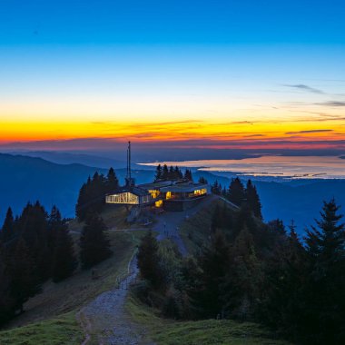 Akşam vakti Bezau Baumgarten teleferiğinin aydınlık dağ istasyonu, dağların üzerinde gün batımı, arka planda gökyüzü ve Constance Gölü turuncu parlıyor.