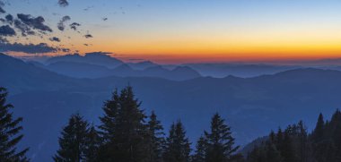 Bregenzerwald panoramasında dağ siluetleri ve arka planda Sntis var. Gün batımı kırmızı ve sisli hatları görünür kılar..