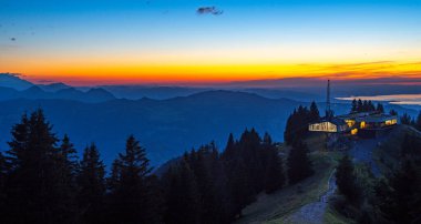 Akşam vakti Bezau Baumgarten teleferiğinin aydınlık dağ istasyonu, dağların üzerinde gün batımı, arka planda gökyüzü ve Constance Gölü turuncu parlıyor.