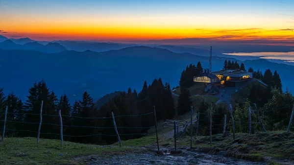 Akşam vakti Bezau Baumgarten teleferiğinin aydınlık dağ istasyonu, dağların üzerinde gün batımı, arka planda gökyüzü ve Constance Gölü turuncu parlıyor.