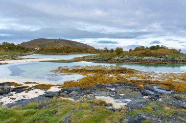 Sommarya, Troms, Norveç 'te taşlarla, yosunlarla ve beyaz kumlarla çevrili mavi turkuaz laguna. Arka planda tatil evi ve dağ var. Macera için özel bir yer  