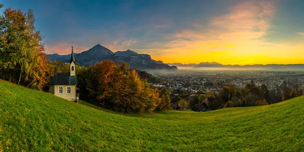 Ren Vadisi 'ndeki Panorama Günbatımı' nda, bir ağaç ve şapel ile, Dornbirn şehrinin üzerinde mavi gökyüzü, çayır ve tarlalar. Arkasında İsviçre dağları var. Artçı, ilginç renkli bulutlar.