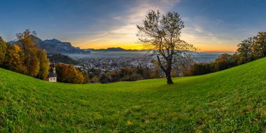 Ren Vadisi 'ndeki Panorama Günbatımı' nda, bir ağaç ve şapel ile, Dornbirn şehrinin üzerinde mavi gökyüzü, çayır ve tarlalar. Arkasında İsviçre dağları var. Artçı, ilginç renkli bulutlar.