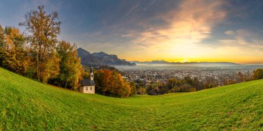 Ren Vadisi 'ndeki Panorama Günbatımı' nda, bir ağaç ve şapel ile, Dornbirn şehrinin üzerinde mavi gökyüzü, çayır ve tarlalar. Arkasında İsviçre dağları var. Artçı, ilginç renkli bulutlar.