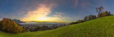 Panorama Sunset in the Rhine Valley, with a tree, blue sky over the city of Dornbirn, meadow and fields. autumn colored trees. beautiful afterglow, interesting colored veil clouds over Swiss mountains clipart