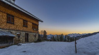 wooden holiday house on hill in afterglow, shingled alpine house on sloppy meadow, next to larch and fir in afternoon mood and view to Bregenz forest. first snow with snowy mountains in background  clipart