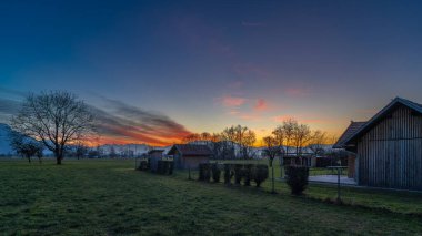 Çayırda ahşap ahır, bahçe ve gün batımının altında baskın ağaç, turuncu, kırmızı bulutlar ve mavi gökyüzü, bahçe alanı, ağaçlar, orman kenarı ve dağ arkaplanı olan otlak manzarası.