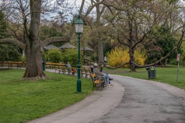 Viyana, Avusturya. 9 Nisan 2023 'te stadtpark (şehir parkı) sakinliğini kucaklarken, insanlar park banklarında dinleniyor.