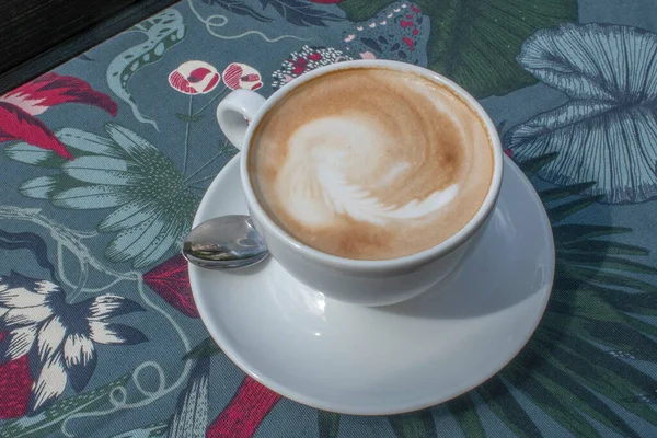 自然の模様が印刷されたテーブルクロスに白いカップで提供されるホットカプチーノのトップダウンビュー 緑豊かな環境の中で心安らぐ飲み物を楽しむ感覚を呼び起こします — ストック写真