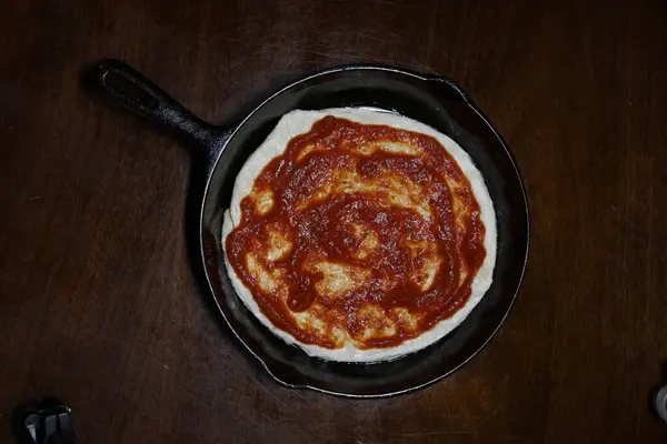 Küçük pizza hamuru dökme demir bir tavaya sıkıştırılmış, pizza sosu her yere yayılmış, koyu ahşap bir masaya yerleştirilmiş. Akşam yemeğinde pizza. İtalyan 00 unundan yapılmış. Akşam yemeği için hazırlanıyorum..