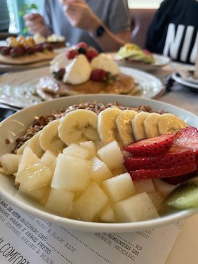 Armut, muz ve çilekli meyve kasesi, arka planda kahvaltı yapan bulanık insanlar.
