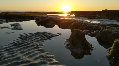 Günbatımında Güney Kaliforniya plaj sahneleri gelgit havuzları, siluetler, soyut kaya oluşumları, okyanus dalgaları, sörf tahtalı sörfçüler ve Swamis Resif Parkı Encinitas California 'da sıcak güneşli gökyüzü.