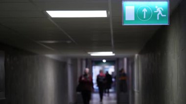 Bulanık şekiller, parlak bir çıkış işaretinin rehberliğinde loş bir koridorda yürür..