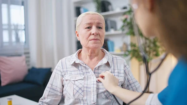 専門の看護師は ホームケアサービスの一環として 彼女の心拍を注意深く聞く古い女性を検査します — ストック写真