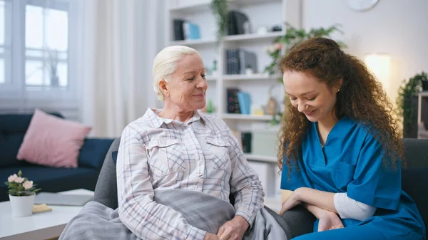 Female nurse keeping company for senior lady at home, women talking and laughing