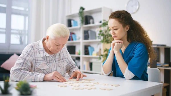 Hemşire yapbozu birleştiren yaşlı kadını izliyor, beyin egzersizi yapıyor.