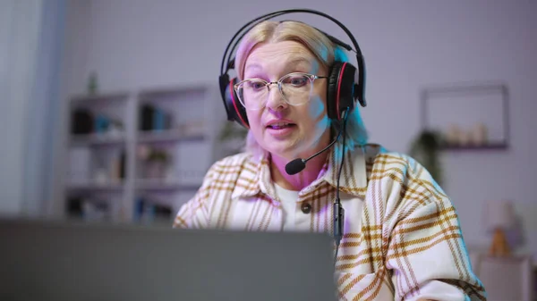 A senior woman in a headset is playing a video game on her laptop as her retirement hobby