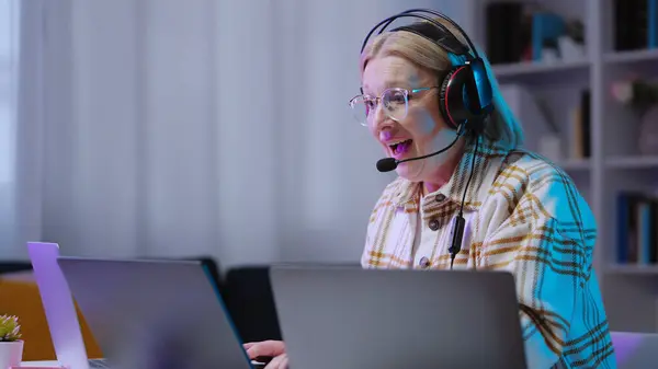 A smiling senior woman gamer in a headset is communicating with other gamers while playing a multiplayer video game