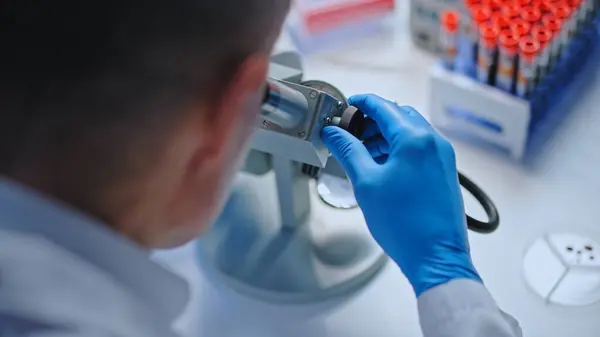 Hospital lab worker studying blood sample under microscope, disease diagnostics