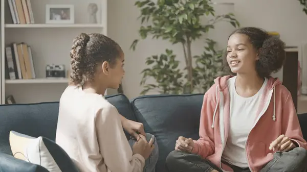 İki siyah genç kız kanepede konuşuyor, arkadaşları evde eğleniyor.