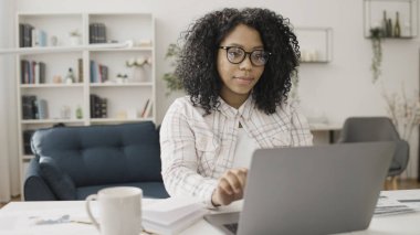 Afro-Amerikalı bir kadın evinde dizüstü bilgisayarıyla çalışıyor ve rahat ofisinde gülümsüyor.
