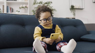 Şık gözlüklü şirin bebek kanepede otururken akıllı telefonuyla oynuyor.