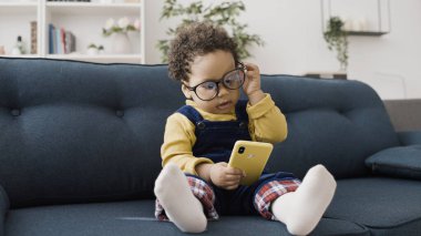 Büyük gözlüklü şirin bir erkek bebek kanepede otururken akıllı telefonuyla oynuyor.