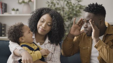 Baba, ağlayan bebeği neşelendirmek için komik suratlar yapıyor. Annenin kucağına oturuyor, ebeveynlik yapıyor.