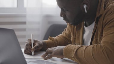 Kulaklıklı Afro-Amerikalı bir adam online ders izliyor ve not alıyor, uzaktan eğitim alıyor.