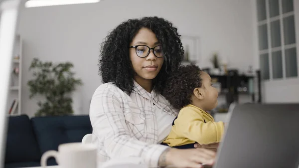 Working mother typing on laptop, baby laughing on her lap, work from home mom
