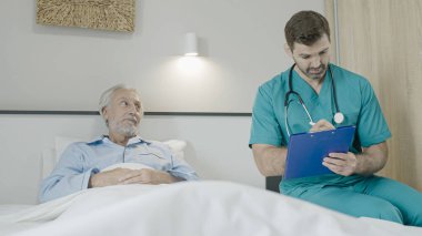 Doktor yatakta kıdemli hastayla konuşuyor, hastanın kartıyla yazıyor, yaşlılara bakıyor.