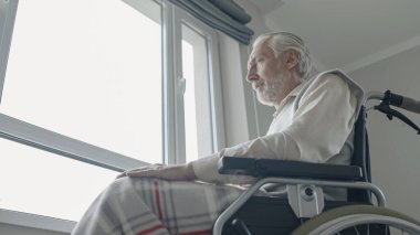 Sakatlığı olan yaşlı adam kendini yalnız hissediyor, pencerenin yanında tekerlekli sandalyede oturuyor.