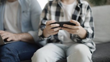 Genç çocuk oyun oynarken babası dizüstü bilgisayarda yazı yazıyor, erkek erkeğe eğleniyor.