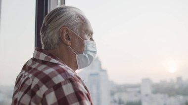 Medikal maskeli üzgün yaşlı adam pencereden dışarı bakıyor, salgın izolasyonu var.