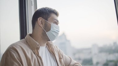Pencereden dışarı bakan koruyucu maskeli orta yaşlı bir adam, salgın güvenliği.