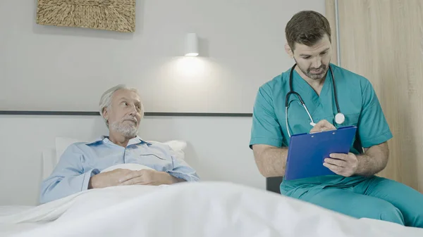 Doctor talking to senior patient in bed, writing down in patient\'s card, elderly care