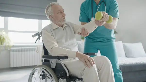 Rehabilitation doctor helping aged man in wheelchair to do exercise with dumbbells