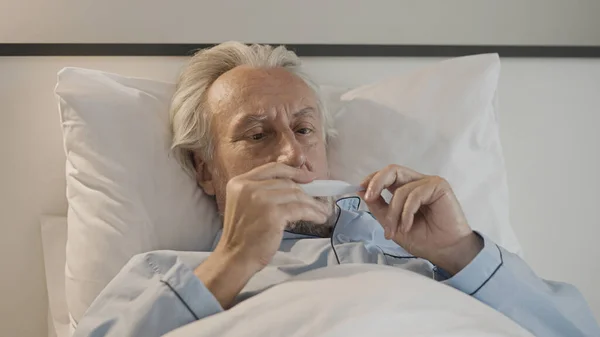 Sad senior man measuring body temperature, lying in bed with viral infection