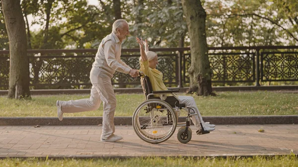 Aktif kıdemli adam, gülümseyen karısı tekerlekli sandalye kullanıcısı ile parkta koşuyor, mutluluk